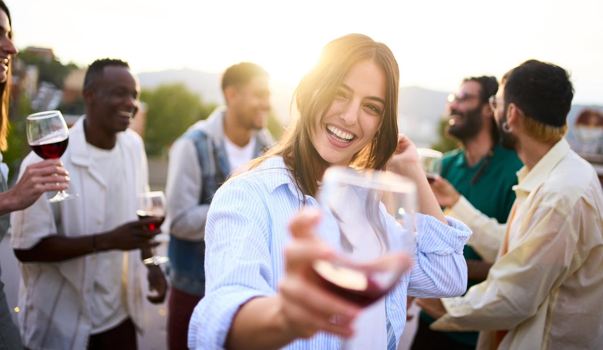 Porträt einer hübschen jungen Frau, die mit einem Glas Rotwein in der Hand in die Kamera schaut und lächelt.