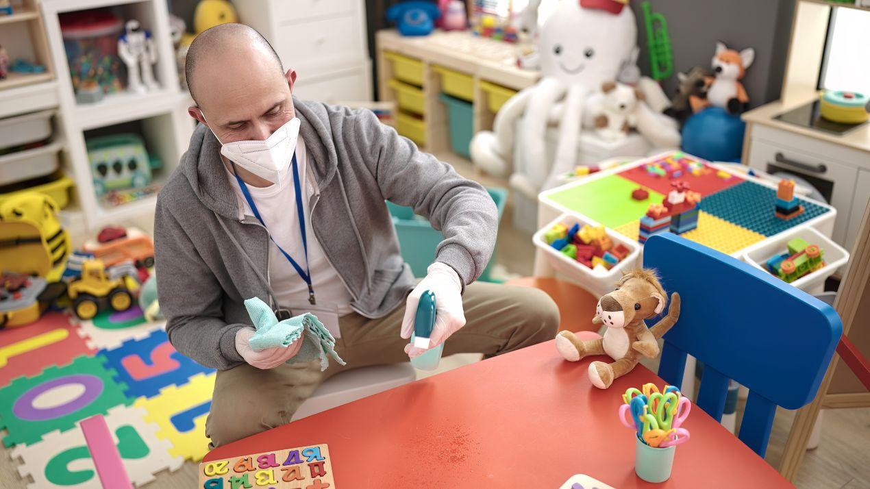 E-Learning Hygiene in Kindertagesstaette: Mann mit Maske desinfiziert Spielzeug zur Praevention von Infektionskrankheiten